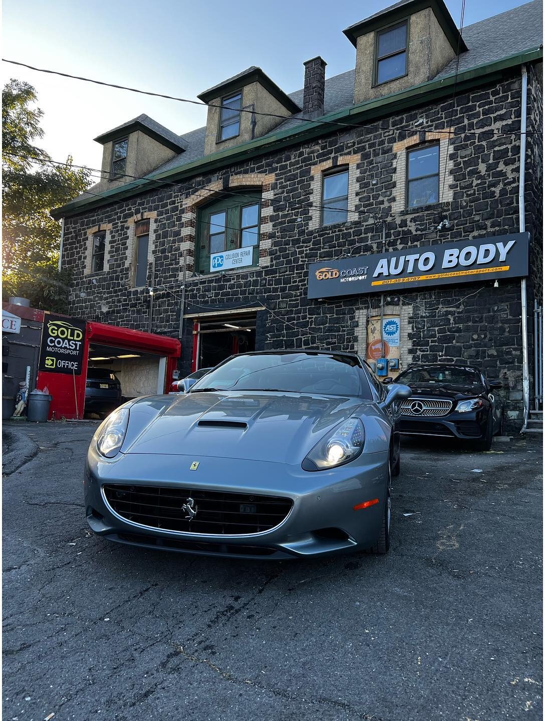 Silver-grey Ferrari California facing the camera in the shop lot, with the Gold Coast Motorsport auto body sign behind it.